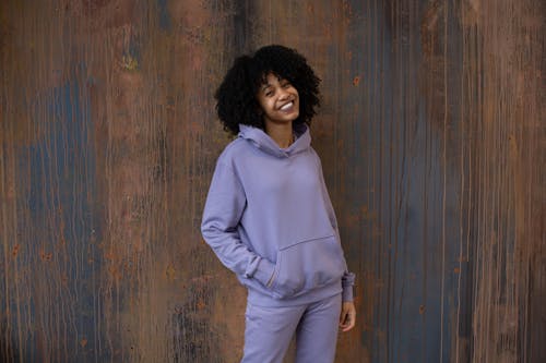 Delighted young African American female with dark curly hair in stylish hooded tracksuit smiling happily and looking at camera while standing against wooden wall