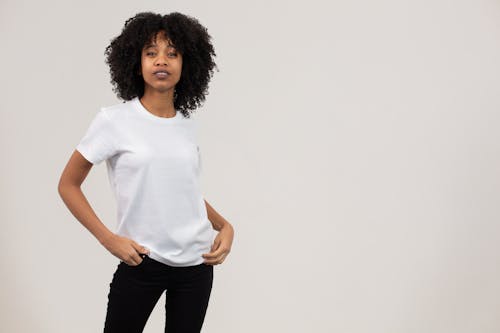 Self esteem young black female model with dark Afro hair in white t shirt standing in light studio with hands in pockets and looking at camera