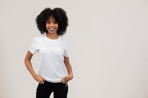 Positive young black female millennial with dark Afro hair in white t shirt smiling and looking at camera while standing in light studio with hands in pockets