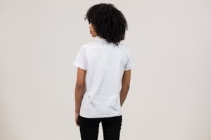 Back view unemotional young African American female wearing casual white t shirt and black pants while standing against light wall