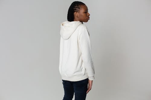 Back view emotionless young African American female in jeans and white comfy hoodie standing against light wall and looking away