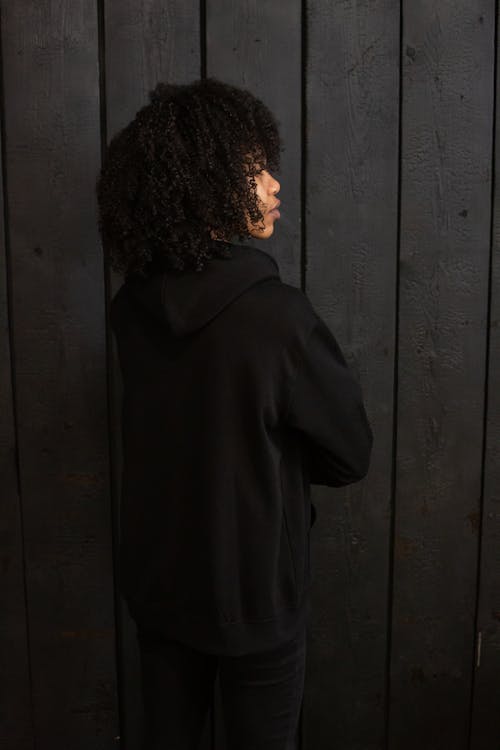 Free Back view emotionless African American female with Afro hairstyle wearing black comfy hoodie standing against dark plank wall Stock Photo