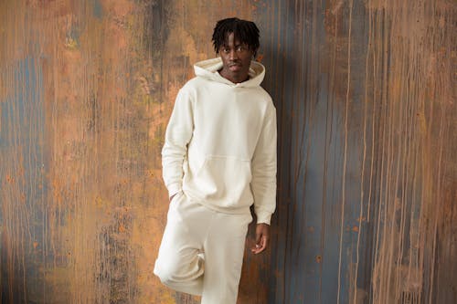 Content African American male in white hoodie and pants standing with hand in pocket near shabby wooden wall and looking at camera