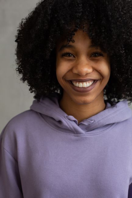 Delighted young African American female with Afro style wearing violet hoodie standing in light room and looking at camera with toothy smile
