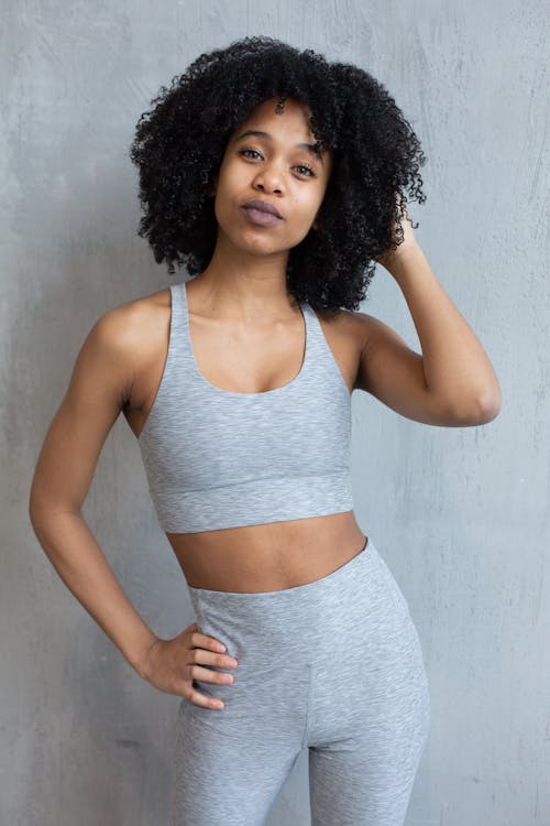 Young fit African American female in sportswear touching curly hair and looking at camera while standing with hand on waist against light wall in studio