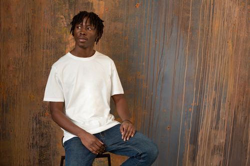 Free Young unemotional male wearing jeans and white t shirt sitting on high stool against wooden wall and looking away Stock Photo