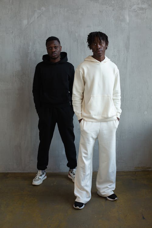 Full body serious African American males in comfy white and black hooded activewear standing with hands in pockets in light studio and looking at camera