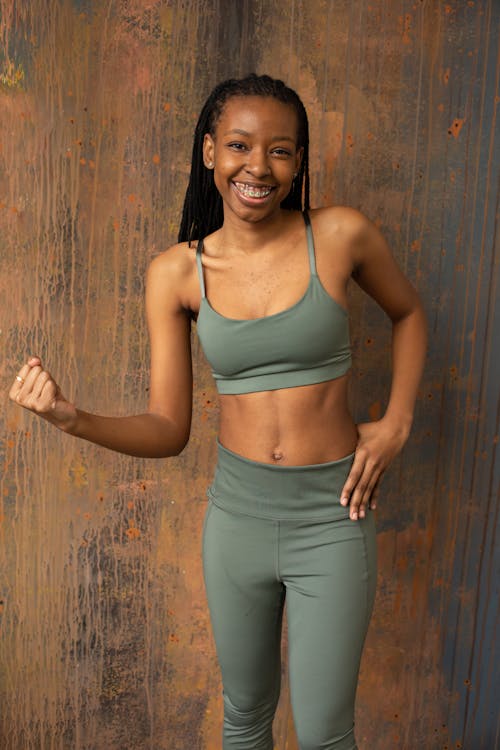 Happy young black lady wearing leggings and sportive bra while looking at camera and standing near brown wall while showing fist in light place