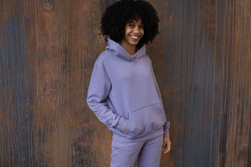 Positive young African American female in purple hoodie and pants standing near brown wall and looking at camera in light place
