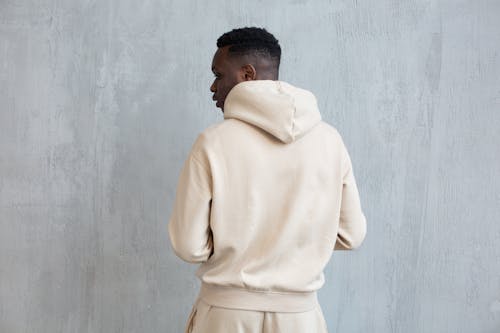 Back view of African American male wearing white hoodie and pants while standing near gray wall in light place