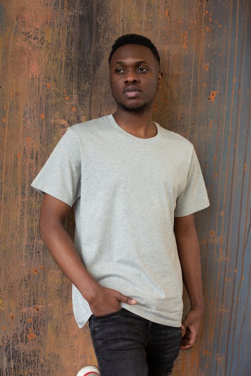 Pensive young black male in casual clothes standing near brown wooden wall in light place and looking away