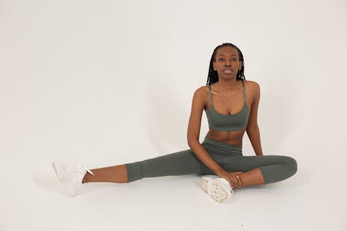 Full body of confident African American lady wearing gray sportive bra and leggings while sitting on floor in bright studio on white background and looking at camera