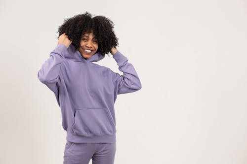 Positive young African American female in purple hoodie and pants standing in light studio on white background and looking at camera