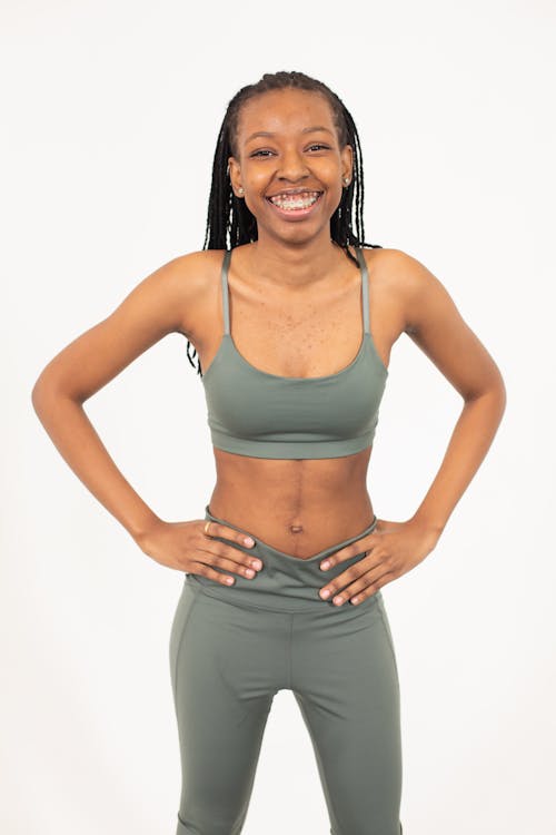 Positive young African American female in sportive bra and leggings standing in light studio on white background and looking at camera