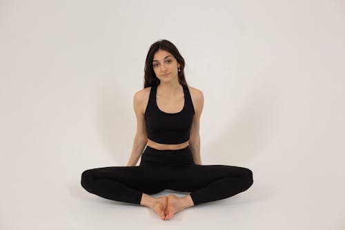 Full body of confident female wearing black sportive bra and leggings while sitting on floor in bright studio on white background and looking at camera