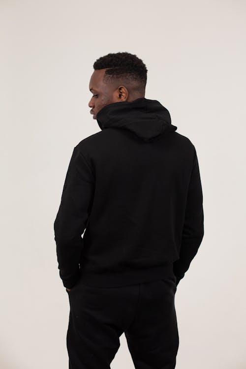 Back view of serious African American male wearing black sportive outfit standing with hands in pockets against white background and looking down