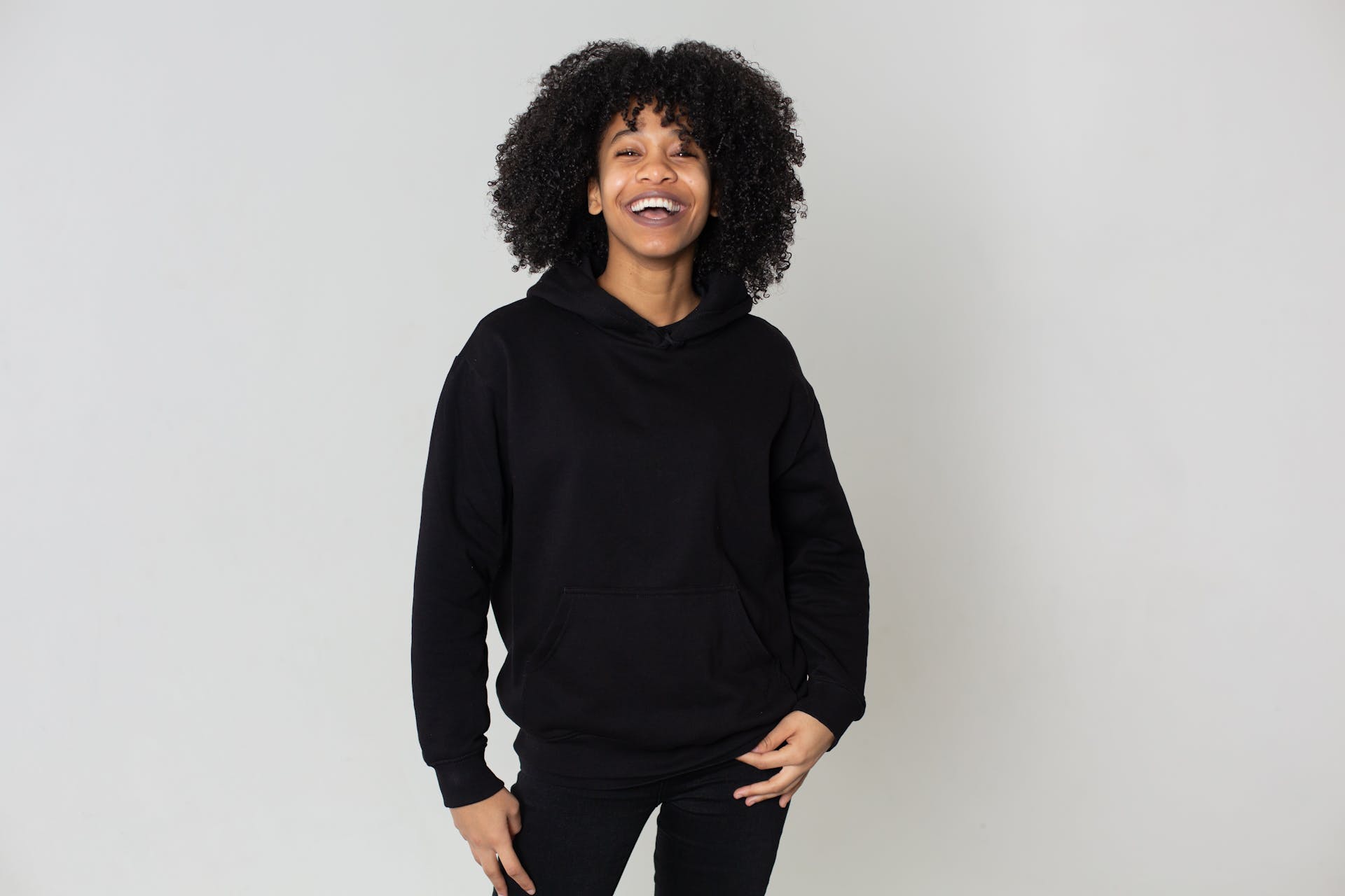 Cheerful African American female with Afro hairstyle wearing trendy black outfit smiling and looking at camera on white background in studio