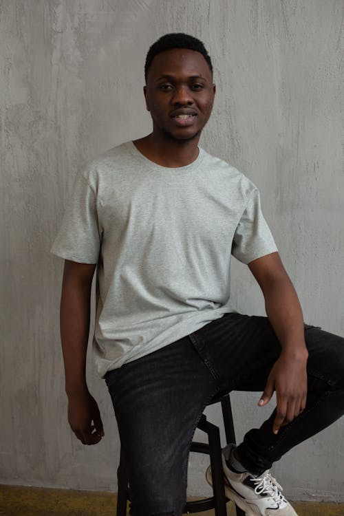 Positive African American male in casual outfit smiling and looking at camera while sitting on chair near wall in studio