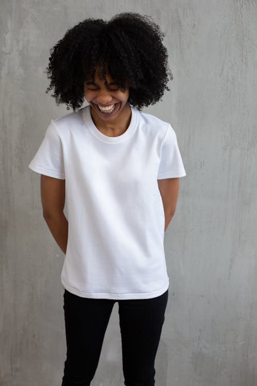 Young African American female in white t shirt with curly hair standing against gray wall and holding hands behind back and laughing happily with closed eyes