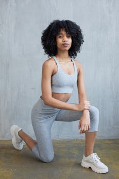 Full body of young African American female in trendy gray top and leggings putting arms on knee in modern studio and looking at camera