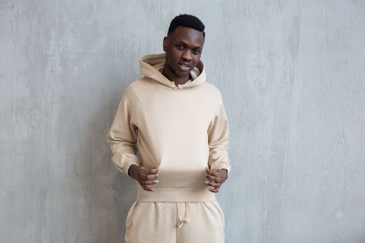 Confident African American guy standing against gray wall and holding hoodie with fingers while looking at camera