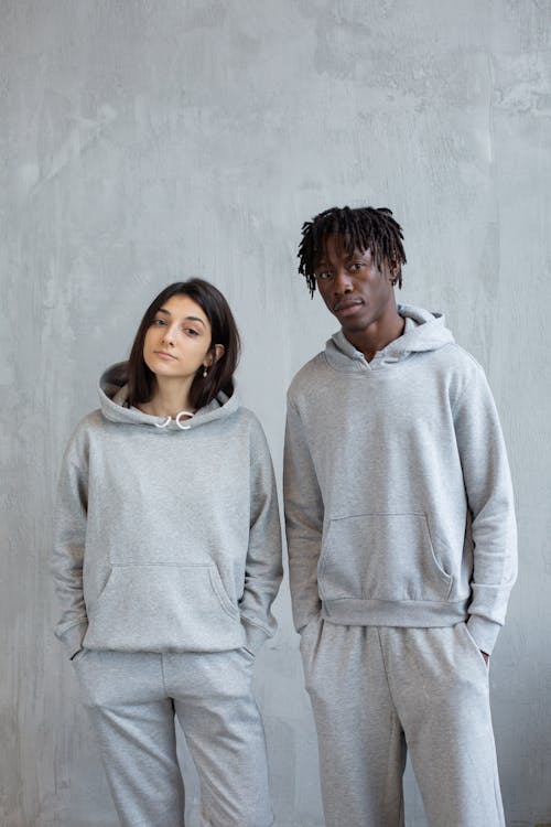 Serious multiracial couple wearing same sportive clothes and standing against concrete wall with hands in pockets