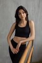 Calm young female athlete in black top and leggings putting elbow on barre while standing against gray wall and looking at camera