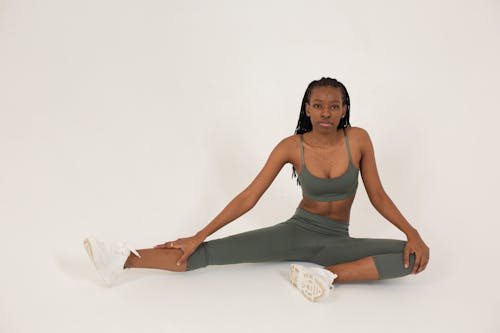 Full body of fit African American female in sportswear sitting on floor against white background and looking at camera