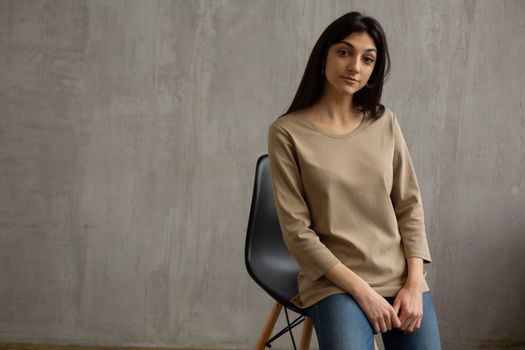 Young female wearing casual outfit sitting on chair and looking at camera against concrete wall