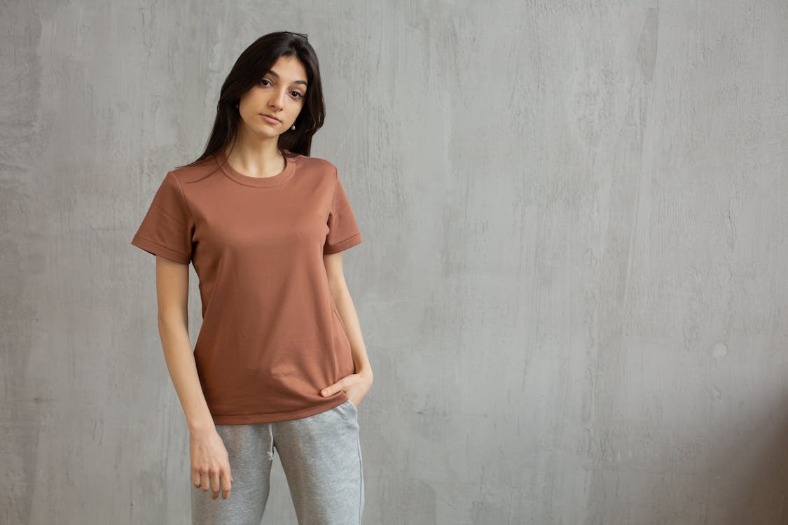 Free Serious female with long hair wearing t shirt and sweatpants standing against gray wall and looking at camera Stock Photo