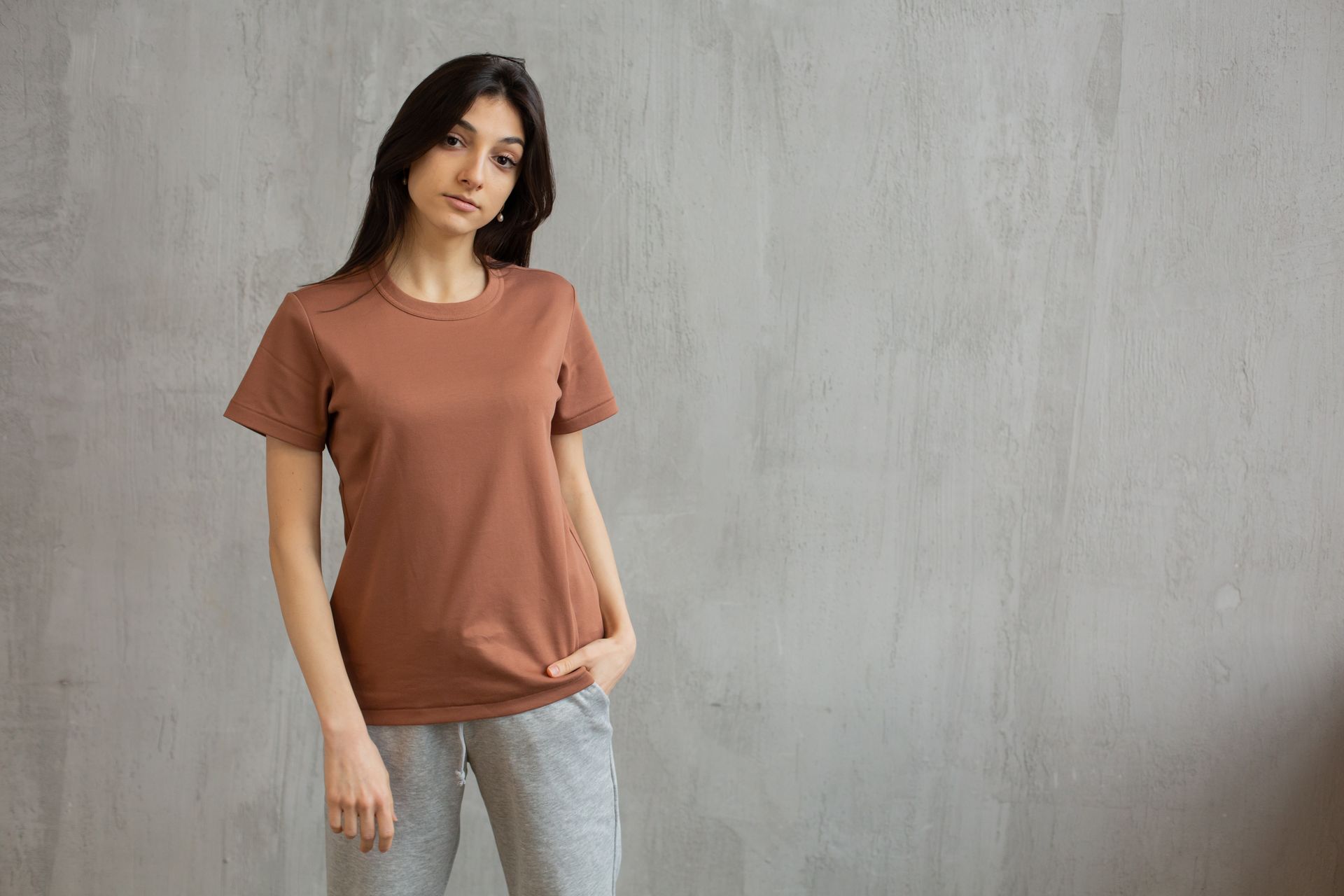 Serious female with long hair wearing t shirt and sweatpants standing against gray wall and looking at camera