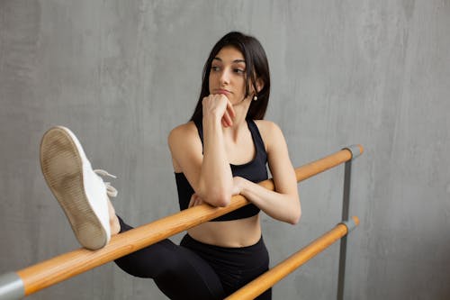 Slim female athlete raising leg on barre while leaning on hand and thoughtfully looking away