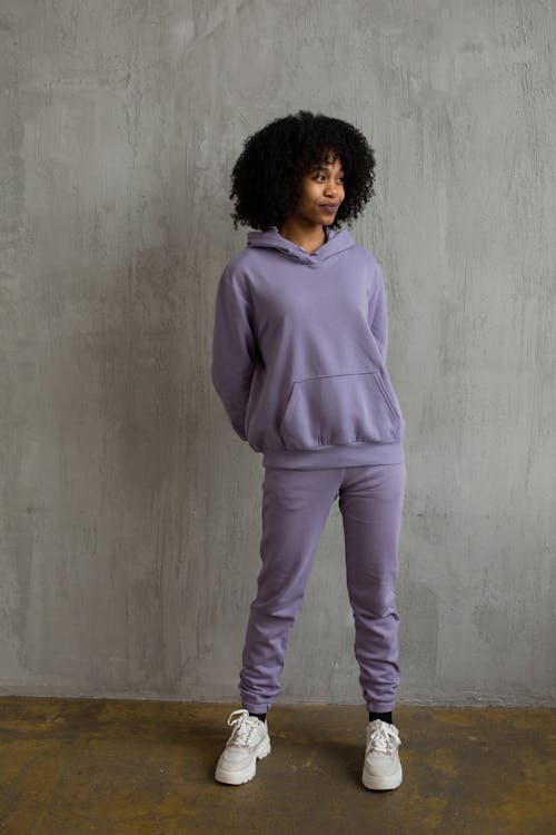 Full body of young African American female with curly hair in trendy violet sportswear holding hands behind back and looking away in modern studio