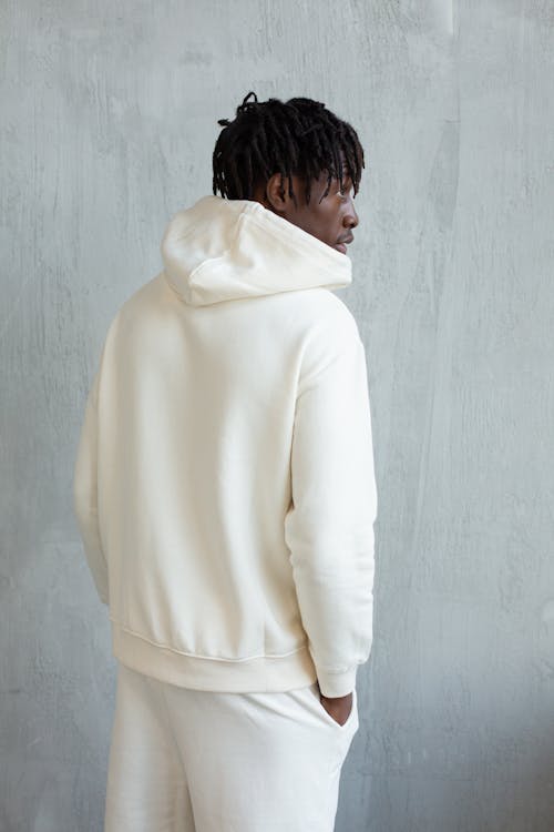 Young African American guy in trendy white hoodie keeping hands in pockets while standing against gray wall and looking away