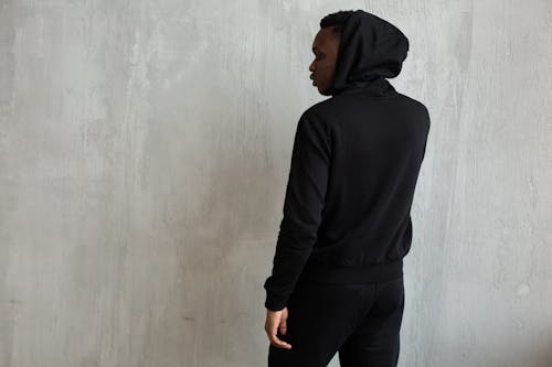 Young African American male in trendy black hoodie with hood on head standing near cement wall and looking away