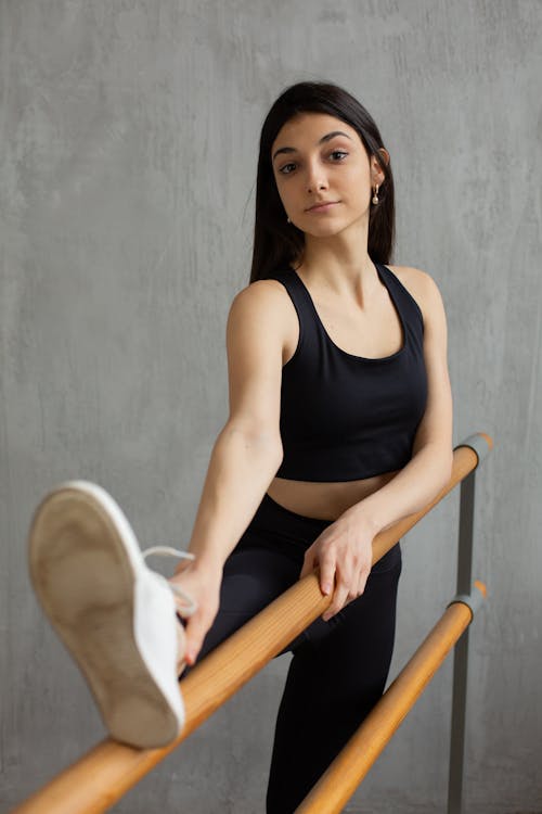 Young slim female with long dark hair in black sportswear stretching legs on barre in ballet studio and looking at camera