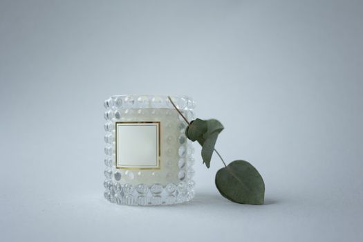 A beautiful glass candle holder with a blank label and eucalyptus leaves against a white background.