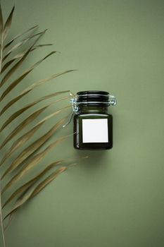 A sleek dark jar with blank label, set beside a tropical leaf on a green backdrop.