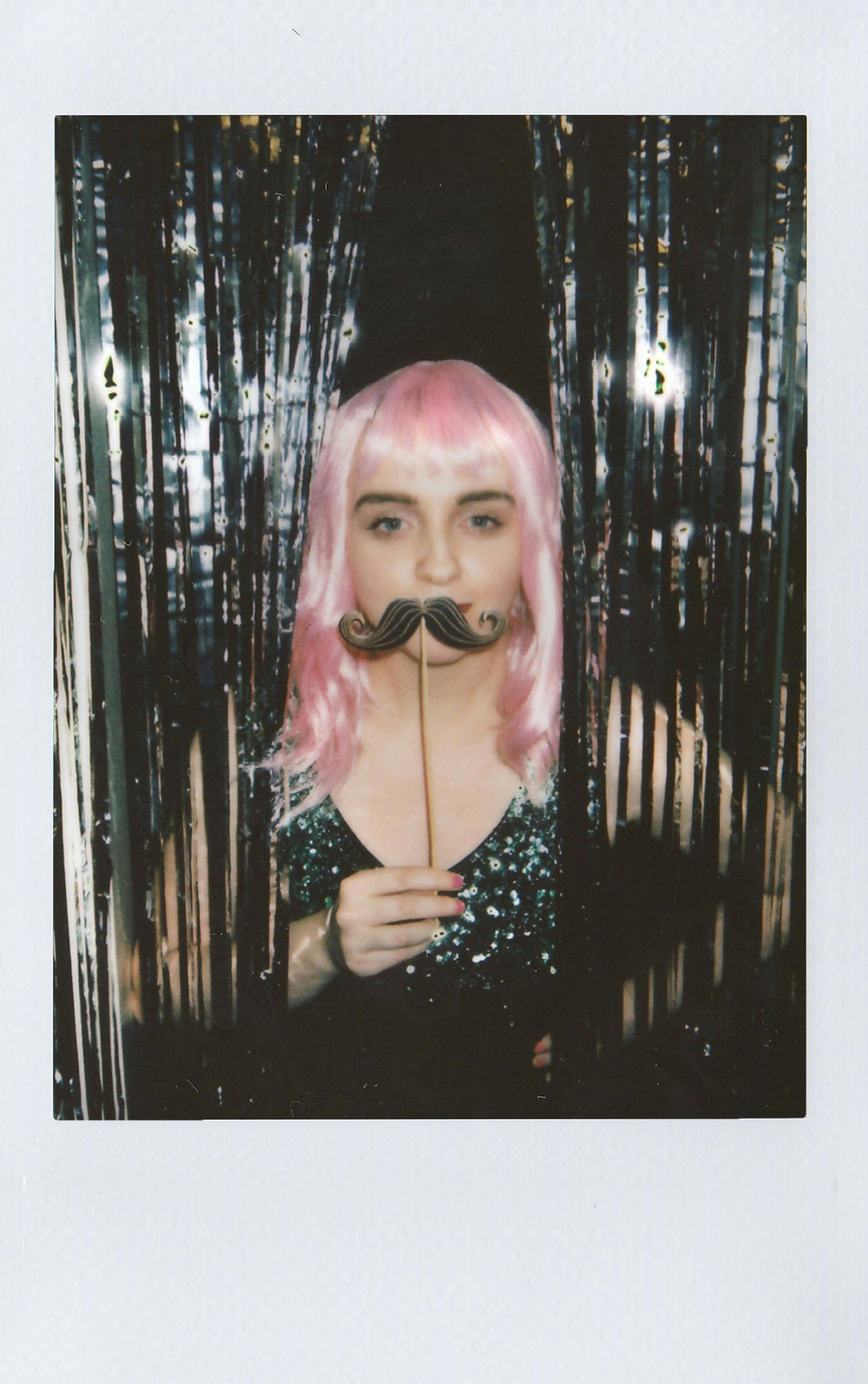 A woman with pink hair poses with a novelty mustache in a sequin top, creating a playful vibe.