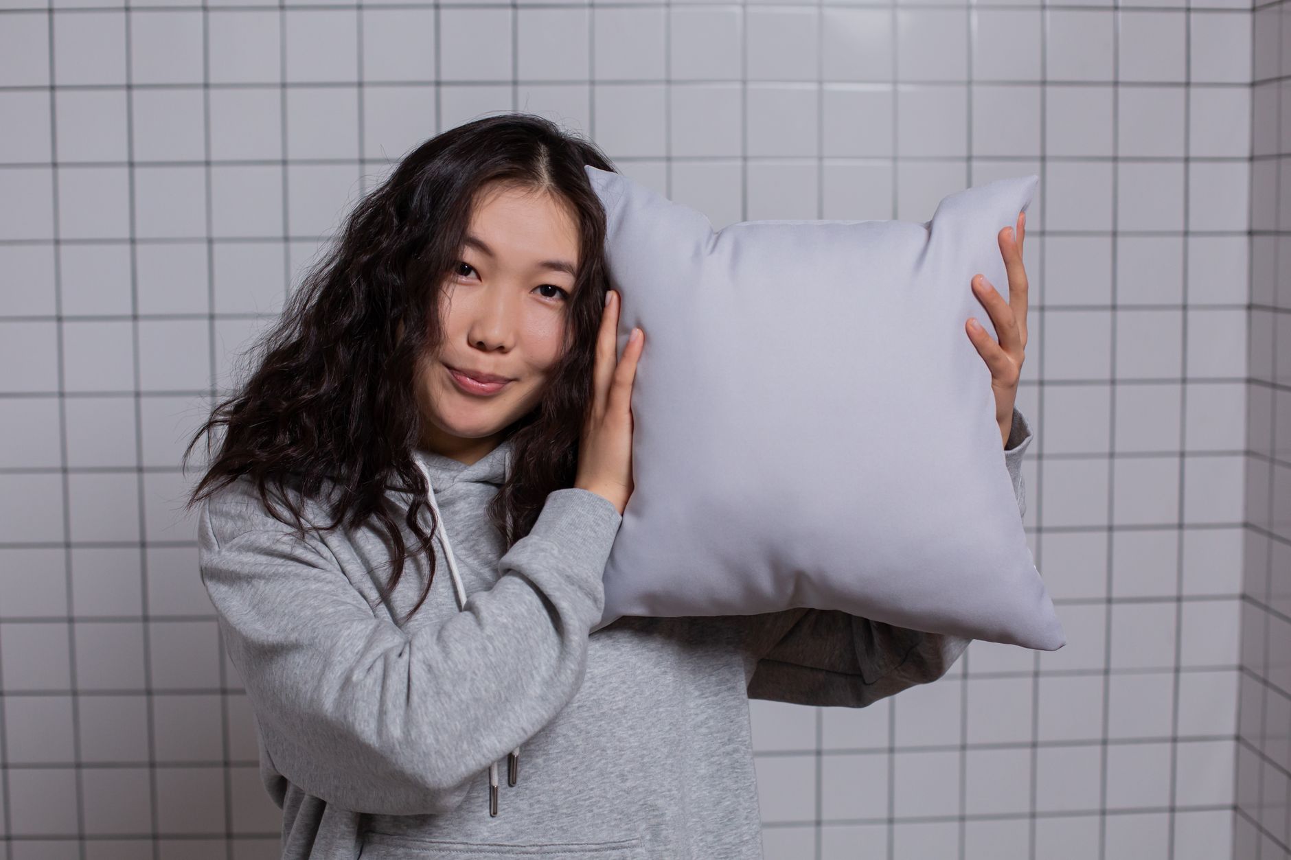 Young woman in a grey hoodie holding a white pillow against a tiled background, indoors.