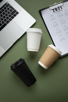 Minimalist flat lay of coffee cups, laptop, and 2021 calendar on green background.