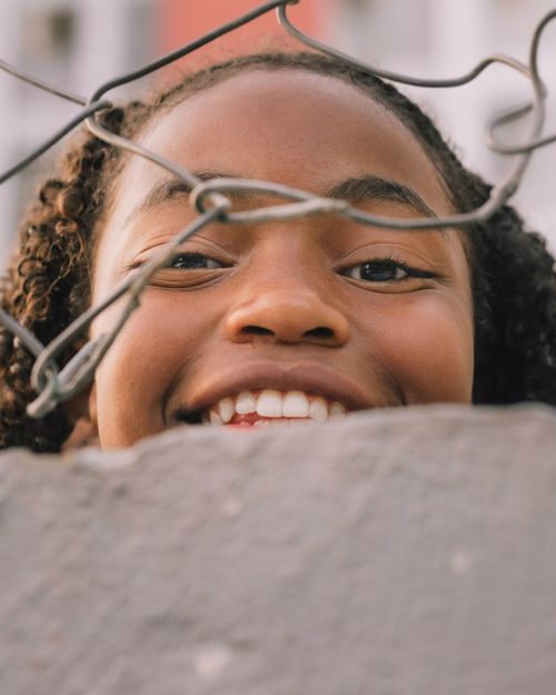 Free Close-up of a happy young girl smiling through a chain link fence outdoors in a warm setting. Stock Photo