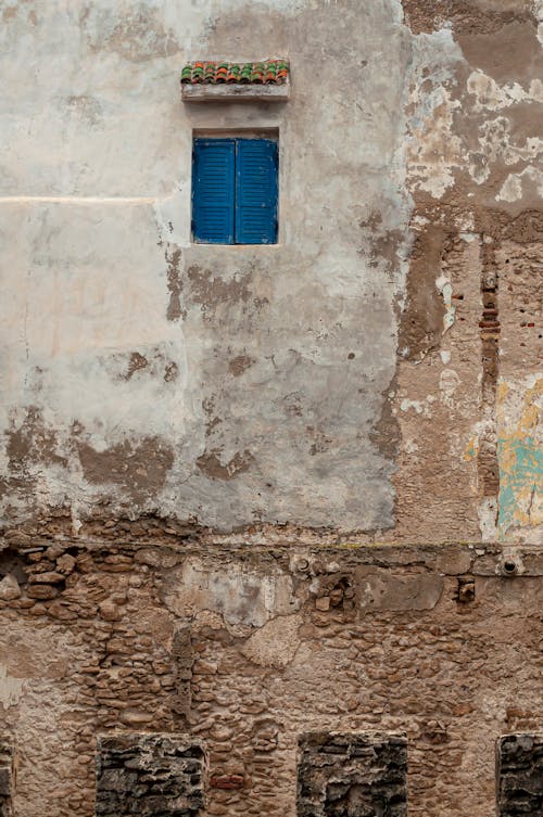 Exterior fragment of rough shabby building with cracked stone wall and blue wooden shutters on window