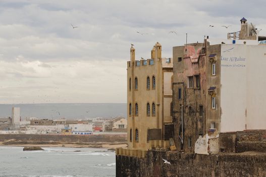 Exterior of aged shabby residential buildings located on embankment over wavy ocean against cloudy sky in Essaouira