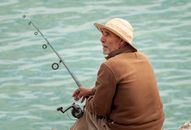 Back view of thoughtful senior ethnic fisherman wearing casual clothed and straw hat while sitting on stone border with rod and looking away