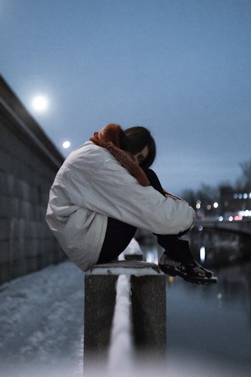 Side view of anonymous female teenager in warm clothes embracing knees on bridge above river in evening in winter