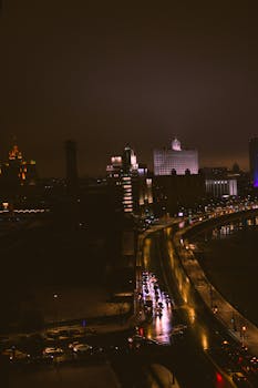 A dramatic view of an urban street at night, showcasing illuminated buildings and reflections on wet pavement.