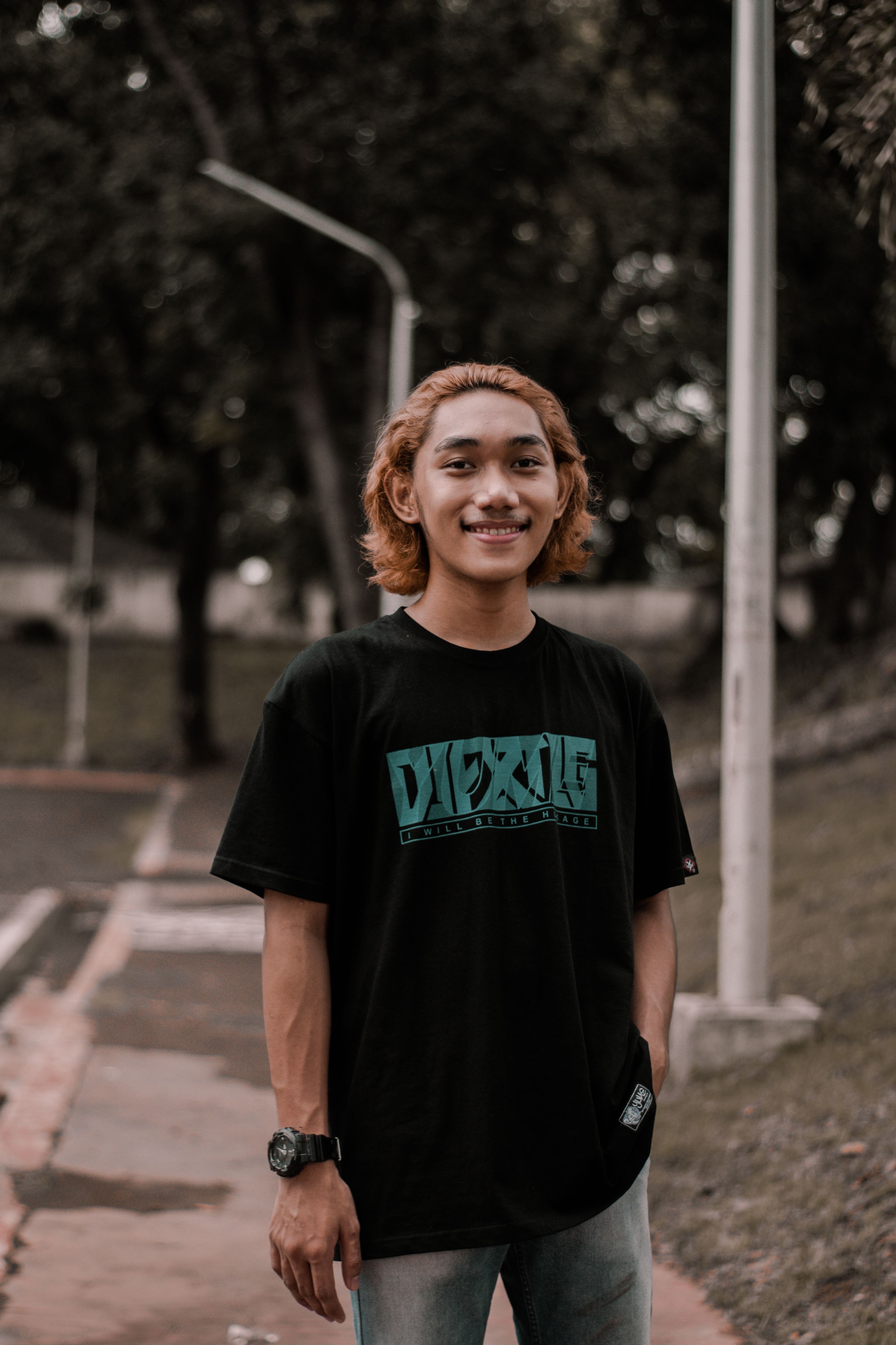 Positive Asian male with dyed hair in casual clothes looking at camera while standing on street with streetlamps in city