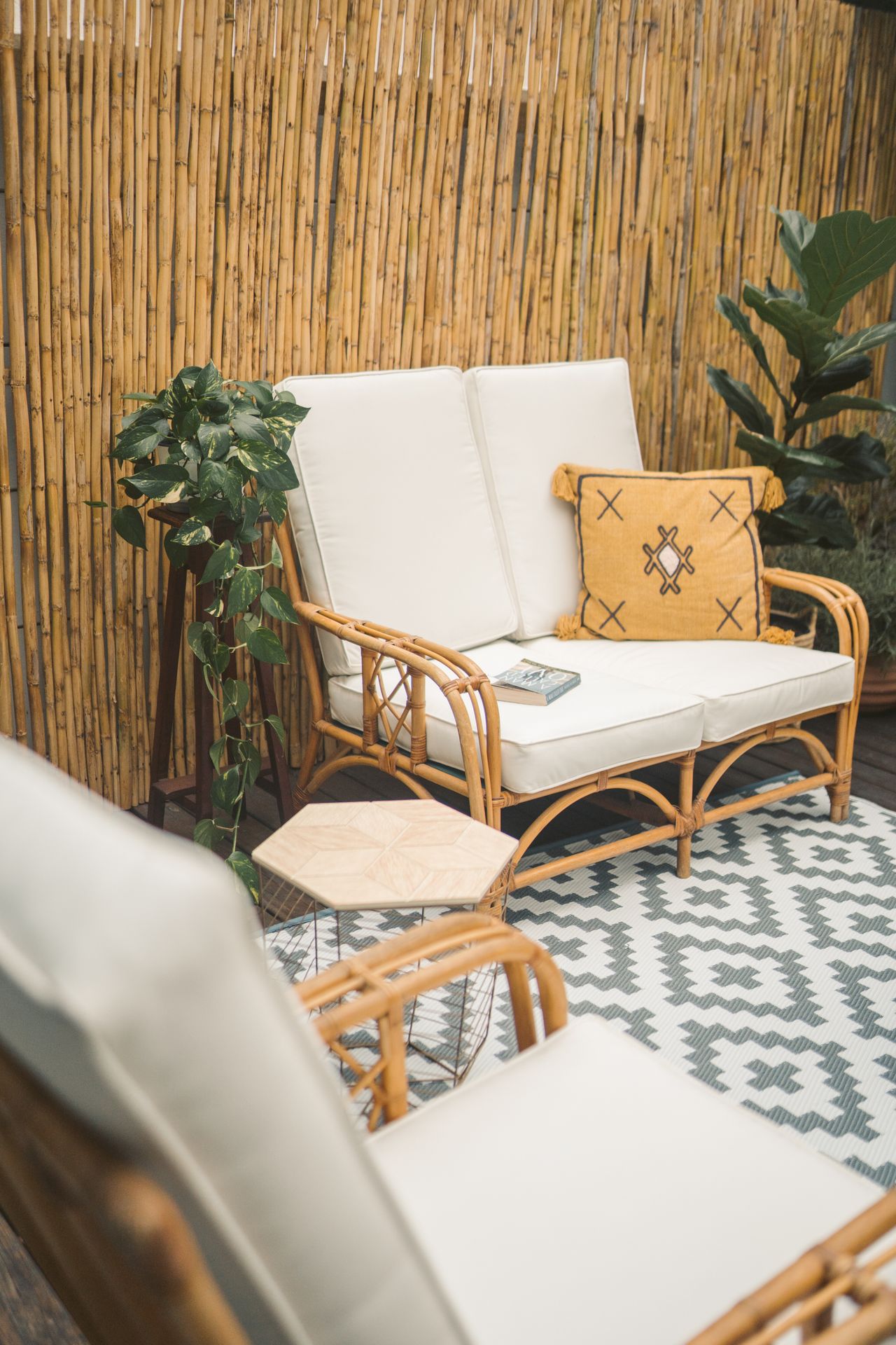 Rattan loveseat with white cushions and a mustard throw pillow on a patterned outdoor rug in a bamboo-screened patio nook with potted plants