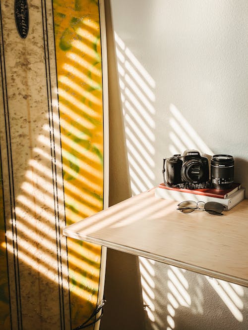 Free From above of photo camera with sunglasses and books laced on wooden table near surfboard leaned on wall Stock Photo